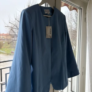 Blå kavaj från NA-KD - Snygg ljusblå kavaj + tillhörande kjol från NA-KD Perfekt för en stilren look. Kavajen har en klassisk design med långa ärmar och en rund halsringning.
