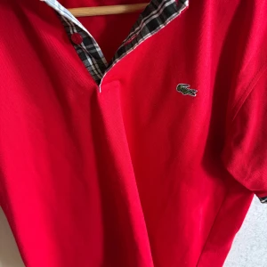Röd pikétröja från Lacoste - Säljer en snygg röd pikétröja från Lacoste med klassisk krage och knappar. Tröjan har ett stilrent rutigt mönster på insidan av kragen och den ikoniska krokodilloggan på bröstet. Perfekt för en sportig och avslappnad look.