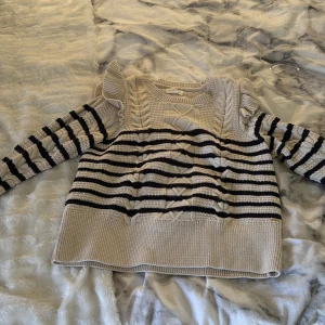 Randig stickad tröja från Holly & Whyte - Säljer en snygg randig stickad tröja från Holly & Whyte. Tröjan har volangdetaljer vid axlarna och ett flätat mönster framtill. Den är beige med svarta ränder och har långa ärmar. Perfekt för en stilren look!