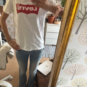 Vit t-shirt från Levi's - Säljer en klassisk vit t-shirt från Levi's med det ikoniska röda logotyptrycket på bröstet. T-shirten är gjord i mjuk bomull och har en normal passform.