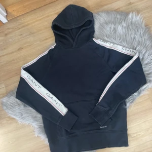 Svart hoodie från GANT - Säljer en stilren svart hoodie från GANT med vita och röda detaljer på ärmarna. Tröjan har en klassisk huva och ribbade muddar. Perfekt för en avslappnad stil. GANT-loggan syns både på ärmarna och framtill.