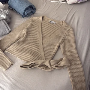 Beige ribbad omlottopp från NA-KD - Säljer en snygg beige ribbad omlottopp från NA-KD. Toppen har långa ärmar och en knytning i midjan som ger en fin passform. Perfekt för en stilren look. Jätte bra skick 