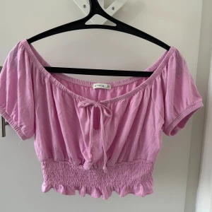 Rosa croppad topp från Cotton On - Söt rosa croppad topp från Cotton On med korta ärmar och en knytning framtill. Toppen har en elastisk smockdetalj vid midjan och en volangkant som ger en feminin touch. Perfekt för varma sommardagar!