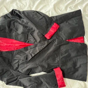 Svart kavaj med röd insida - Snygg svart kavaj med en iögonfallande röd insida. Kavajen har en klassisk design med knappar framtill och långa ärmar. Perfekt för att ge en färgklick till din outfit.