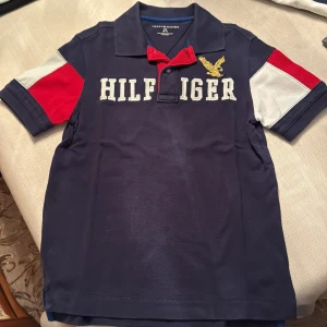 Blå kortärmad pikétröja från Tommy Hilfiger - Säljer en blå kortärmad pikétröja från Tommy Hilfiger med broderad gul örn och stora bokstäver på framsidan. Tröjan har färgblock i rött och vitt på ärmarna och en klassisk krage med knappar. Perfekt för en stilren look!
