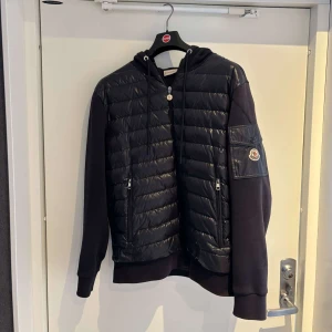 Svart jacka med huva från Moncler - Snygg marinblå jacka från Moncler med quiltad framsida och dragkedja. Jackan har en huva och en ficka med knapp på ärmen. Perfekt för kyligare dagar. Storleken är XL men passar M/L