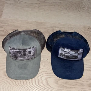 Två kepsar med bilmotiv - Säljer två kepsar från Gumball 3000. Den ena är grå med ett svartvitt bilmotiv och den andra är blå med ett liknande motiv. Båda kepsarna har mesh på baksidan och justerbar passform.