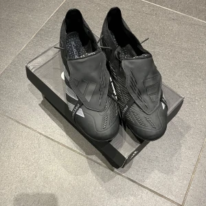 Adidas Predator  - Snygga svarta Adidas Predator fotbollsskor med stilrena vita detaljer. Skorna har en modern design med texten 'EST. 1994' och är perfekta för fotbollsplanen. De är tillverkade i syntetmaterial för optimal prestanda.