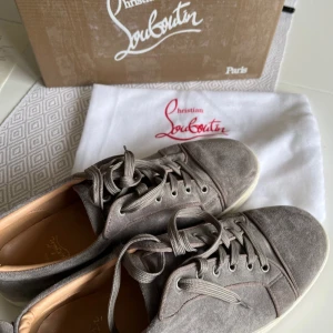 Grå sneakers från Christian Louboutin - Snygga grå sneakers från Christian Louboutin med den ikoniska röda sulan. Skorna har snörning och en rund tå, vilket ger en klassisk och stilren look. Perfekta för dig som vill ha en touch av lyx i vardagen.