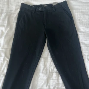Svarta chinos från Jack & Jones - Snygga svarta chinos från Jack & Jones i modellen Slim/Marco. De har en slim passform och är perfekta för en stilren look. Byxorna har knappar och dragkedja framtill samt fickor både fram och bak.