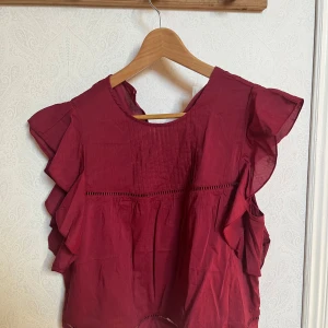 Vinröd blus med volangdetaljer - Snygg vinröd blus med volangdetaljer på ärmarna. Blusen har en rund halsringning och dekorativa sömmar framtill. Perfekt för en stilren look.