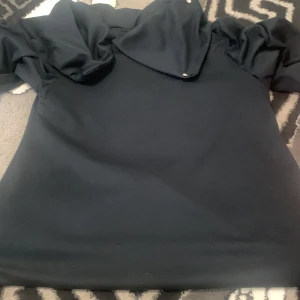 Svart tröja med volangdetaljer - Snygg svart tröja med unika volangdetaljer på axlarna. Perfekt för att ge en stilfull touch till din outfit. Tröjan har en hög krage med knappar som ger en elegant look.