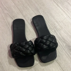 Svarta sandaler med flätad detalj - Snygga svarta sandaler med en bred flätad rem över foten. Perfekta för en stilren look under varma dagar. De har en fyrkantig tå och är tillverkade i syntetmaterial.