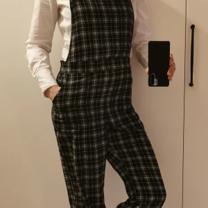 Svartvit rutig byxdress - Snygg byxdress från Monki med svartvitt rutmönster och hängslen. Perfekt för en stilren look. Passar bra med en enkel topp under om man bar vill slänga på sig något snabbt men ändå snyggt. Storlek 38. Längd: 129 cm (ej banden inräknade). Justerbara band. 