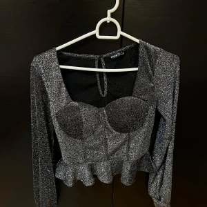 Glittrig svart topp med volang - Snygg glittrig svart topp med långa ärmar och volangdetalj i nederkant. Toppen har en korsettliknande design framtill och knytning i nacken. Perfekt för festliga tillfällen!