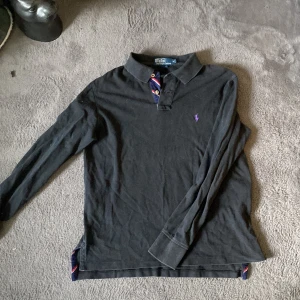 Svart långärmad pikétröja från Ralph Lauren - Säljer en stilren svart långärmad pikétröja från Ralph Lauren med en liten lila logga på bröstet. Tröjan har en klassisk krage och knappar i halsen. Perfekt för en avslappnad men ändå snygg look.