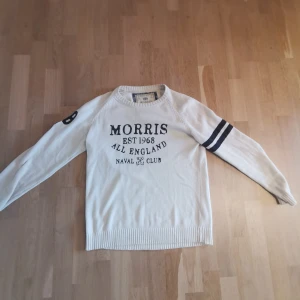 Vit tröja från Morris - Säljer en stilren vit tröja från Morris med tryck på framsidan och två mörka ränder på ena ärmen. Tröjan är tillverkad i 100% bomull och har långa ärmar med ribbstickade muddar. Perfekt för en avslappnad stil.