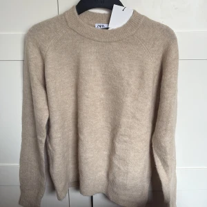 Beige stickad tröja från Zara - Säljer en stilren beige stickad tröja från Zara. Tröjan har en klassisk rund hals och långa ärmar, perfekt för kyligare dagar. Den är mjuk och bekväm, vilket gör den till ett mångsidigt tillskott i garderoben. Passar bra till både jeans och kjol.