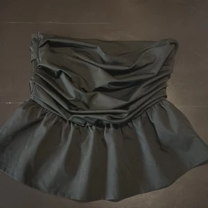 Svart axelbandslös topp - Snygg svart axelbandslös topp med draperad design och volangdetaljer. Perfekt för en stilren och elegant look. Passar bra till både jeans och kjol.