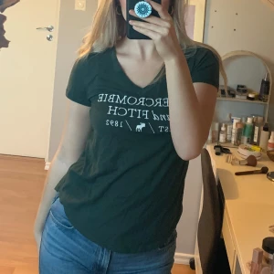 Grön t-shirt från Abercrombie & Fitch - Säljer en snygg grön t-shirt från Abercrombie & Fitch med vit text och logga på framsidan. T-shirten har en v-ringad hals och korta ärmar, perfekt för en avslappnad stil.
