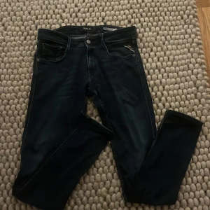Replay Anbass mörkblå jeans - Säljer ett par snygga Replay Anbass jeans i mörkblå tvätt. Klassisk femficksmodell med raka ben och läderpatch bak i midjan. Jeansen har diskreta slitningar och silverfärgade detaljer. Perfekta för dig som gillar stilrena och moderna jeans.