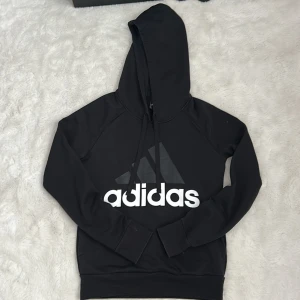 Svart Adidas hoodie med logga XS - Svart hoodie från Adidas i storlek XS med stor vit logga framtill och liten logga bak. Klassisk huva med snörning och långa ärmar. Tillverkad i mjuk bomullsblandning, perfekt för chill eller träning. Snygg och enkel stil som funkar året runt.