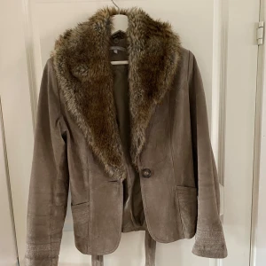 Mockajacka med fuskpäls - Snygg beige mockajacka från FRN by fansa med bred krage i fuskpäls. Jackan har knäppning framtill, två fickor och bälte i midjan. Perfekt för dig som vill ha en stilren och trendig look med lite edge. Passar till både jeans och klänning.