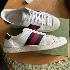 Gucci ace  - Snygga vita sneakers från Gucci med klassiska röd- och blå ränder på sidorna. Skorna har svart detalj i ormskinnsmönster på hälen med Gucci-logga. Tillverkade i skinn med snörning och platt sula. Perfekta för dig som vill ha en lyxig och stilren look. Dessa är självklart äkta och är rätt så sällsynta. Skicket är bra och nypriset ligger på 8000. De passar storlek 42 