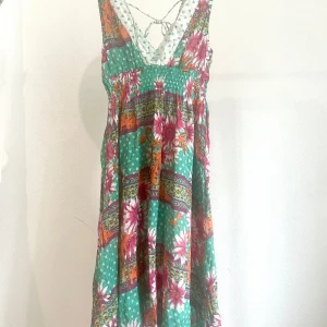 Grön blommig långklänning med knyt i ryggen - Säljer en färgglad långklänning med grönt, vitt och rosa blommönster samt orange detaljer. Klänningen har djup v-ringning och knytning i ryggen. Passar storlek XS-S.