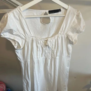 Vit blus med puffärmar från KappAhl - Söt vit blus från KappAhl med korta puffärmar och rund halsringning. Dekorativa veck framtill och liten knytning vid bysten. Perfekt till sommaren för en fräsch look.