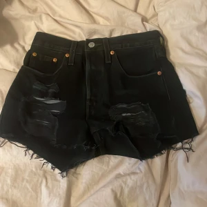 Svarta Levi's jeansshorts med slitningar - Svarta jeansshorts från Levi's med hög midja, råa fransiga kanter och slitna detaljer framtill. Klassisk femficksmodell med Levi's-patch bak och knappgylf. Perfekta för en edgy och avslappnad stil.