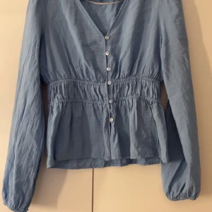 Blå blus med knappar från Vero Moda - Supersöt ljusblå blus från Vero Moda i storlek XS. Blusen har v-ringning, knappar framtill och smockad midja som ger en snygg siluett. Långa ärmar med resår vid handlederna. Perfekt för vår och sommar!