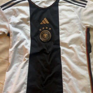 Tysklands matchtröja S - Säljer en officiell Tyskland fotbollströja från Adidas med klassisk svart mittremsa och vita sidor. Tröjan har guldiga detaljer, fyra stjärnor ovanför klubbmärket och svarta Adidas-ränder på axlarna. Materialet är lätt och andas, perfekt för fotboll eller supporterliv.