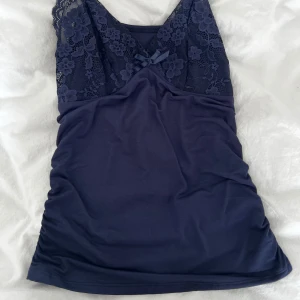 Mörkblå spetsdetaljerad linnetopp - Supersöt mörkblå linnetopp med tunna axelband och v-ringning. Överdelen har blommig spets och en liten rosett framtill. Den har justerbara band. 