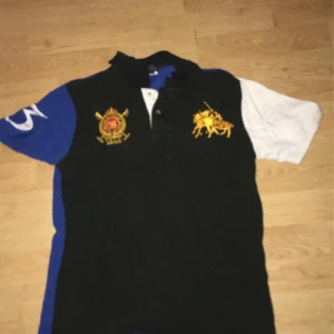 Svart pikétröja med brodyr från Ralph Lauren  - Säljer en svart pikétröja från Ralph Lauren med broderade detaljer på bröstet. Ena ärmen är blå med en vit siffra, den andra är vit. Klassisk krage och knappar framtill.