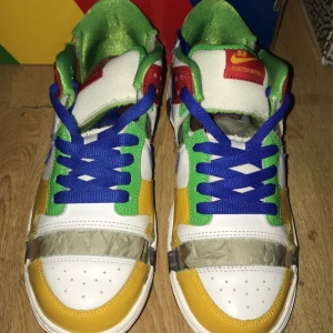 Nike SB Dunk Low eBay collab sneakers i färgglad design - Nike SB Dunk Low eBay collab sneakers med vit bas och detaljer i gult, grönt, blått och rött. Skorna har blå snörning, grön insida och SB-logga på hälen. Klassisk låg modell med rund tå och platt sula. Kommer i originalkartong med färgglad design.