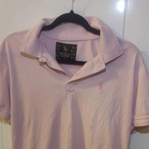 Ljusrosa pikétröja från Polo Ralph Lauren - Ljusrosa pikétröja med klassisk krage och korta ärmar från Polo Ralph Lauren. Tröjan har en broderad logga på bröstet och är tillverkad i mjuk bomull. Perfekt för en avslappnad och stilren look.
