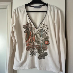 Vit långärmad topp med blommigt tryck från KappAhl - Säljer en vit långärmad topp från KappAhl med v-ringning och dekorativ brun spetskant. 