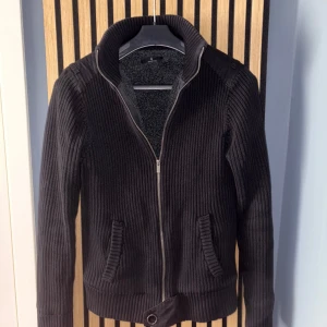 Marinblå stickad jacka från Selected - Säljer en marinblå stickad jacka från Selected med dragkedja och mysigt teddyfoder på insidan. Jackan har ribbade detaljer, hög krage och två fickor framtill. Perfekt för kyliga dagar.