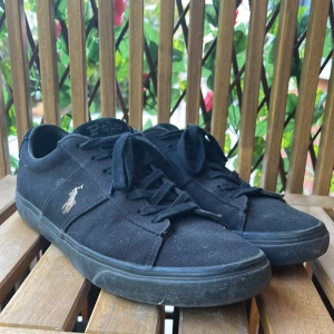 Svarta sneakers från Polo Ralph Lauren - Svarta sneakers från Polo Ralph Lauren med klassisk logga på sidan. Skorna har snörning, rund tå och platt sula. Ovandelen är i textil och sulan i gummi. Tyget på vänstra sko sulan är lite lös men märks inte mycket vid användning.
