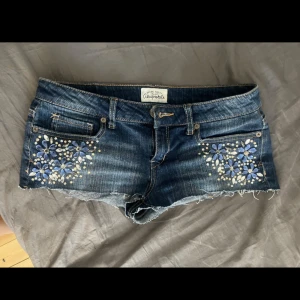 Jeansshorts med blommiga stenar från Aeropostale - Mörkblå jeansshorts med råa kanter och dekorativa blommor av blå och silvriga stenar framtill. Klassisk femficksmodell med låg midja och snygga slitningar. Perfekta för sommardagar när du vill sticka ut lite extra.