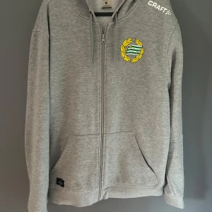 Grå zip-hoodie  Hammarby  - Grå hoodie med dragkedja från Craft. Tröjan har huva med dragsko, två fickor fram och en broderad logga på bröstet. Perfekt för chill eller träning. Snygg och enkel design med vit Craft-text på axeln.