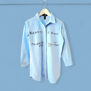 Blå Beastie Boys skjorta med tryck - Ljusblå skjorta från Suzanne Betro med svart Beastie Boys-tryck och texten 'Frozen Metal Haze' både fram och bak. Skjortan har klassisk krage, knappar framtill och långa ärmar. Materialet är bomull och modellen är rak och lite loose.