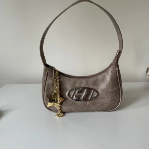 Beige axelväska med metallogo - Snygg beige axelväska i skinnimitation med stort H! metallogo framtill. Väskan har dragkedja upptill och dekoreras med en guldfärgad stjärnberlock och en glittrig hjärtberlock. Perfekt storlek för det viktigaste och ger en cool 00-talsvibe.
