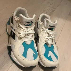 Reebok sneakers vit och turkos - Snygga Reebok sneakers i vitt med turkosa och svarta detaljer. Skorna har klassisk snörning, rund tå och platt sula. Ovandelen är i textil och syntet med cool retrostil och Reebok-logga på sidan och plösen. Perfekta för dig som gillar streetwear och sportig vibe.