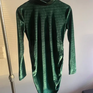 Grön sammetsklänning med lång ärm - Mörkgrön sammetsklänning med lång ärm och rak passform. Klänningen har en enkel design utan synliga detaljer och slutar strax ovanför knäna. Perfekt för dig som gillar en stilren look med lite lyxig känsla.