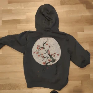 Mörkgrå hoodie med körsbärsblom-tryck - Säljer en mörkgrå hoodie med dragkedja och huva. På ryggen finns ett stort tryck av en gren med rosa körsbärsblommor mot en vit cirkel.