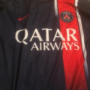 PSG fotbollströja Mbappé 7 - Säljer en officiell Paris Saint-Germain fotbollströja med Mbappé 7 på ryggen. Tröjan är mörkblå med röd och vit detalj på framsidan, Qatar Airways som sponsor och PSG-logga på bröstet. Perfekt för fans av Mbappé och PSG.