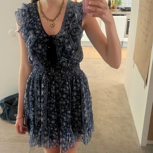 Blå blommig volangklänning - Supersöt blå kortklänning med blommigt mönster och volangdetaljer längs axlar och byst. Klänningen har v-ringning och är ärmlös, med smockad midja som ger en fin passform. Perfekt för sommarens alla tillfällen! 🩷🩷 Den är i nyskick utan prislapp. 