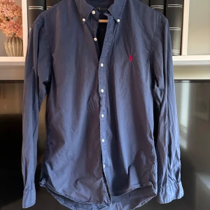 Blå skjorta från Ralph Lauren - Klassisk blå skjorta från Ralph Lauren med knappar hela vägen och button-down krage. Liten röd broderad logga på bröstet. Perfekt till jeans eller chinos för en stilren look. Skriv för frågor och funderingar!!